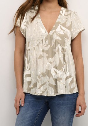 Kobieta ubrana w bluzkę z krótkim rękawem w beżowo-biały wzór z dekoltem w szpic, połączoną z niebieskimi dżinsami, stojąca na jasnym tle.