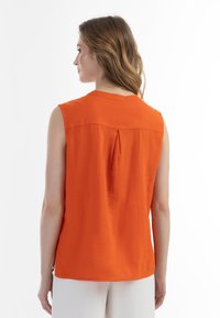 Ærmeløs orange bluse med en rygfælde, rund halsudskæring og sideslids. lavet af let stof, der har en glat tekstur.