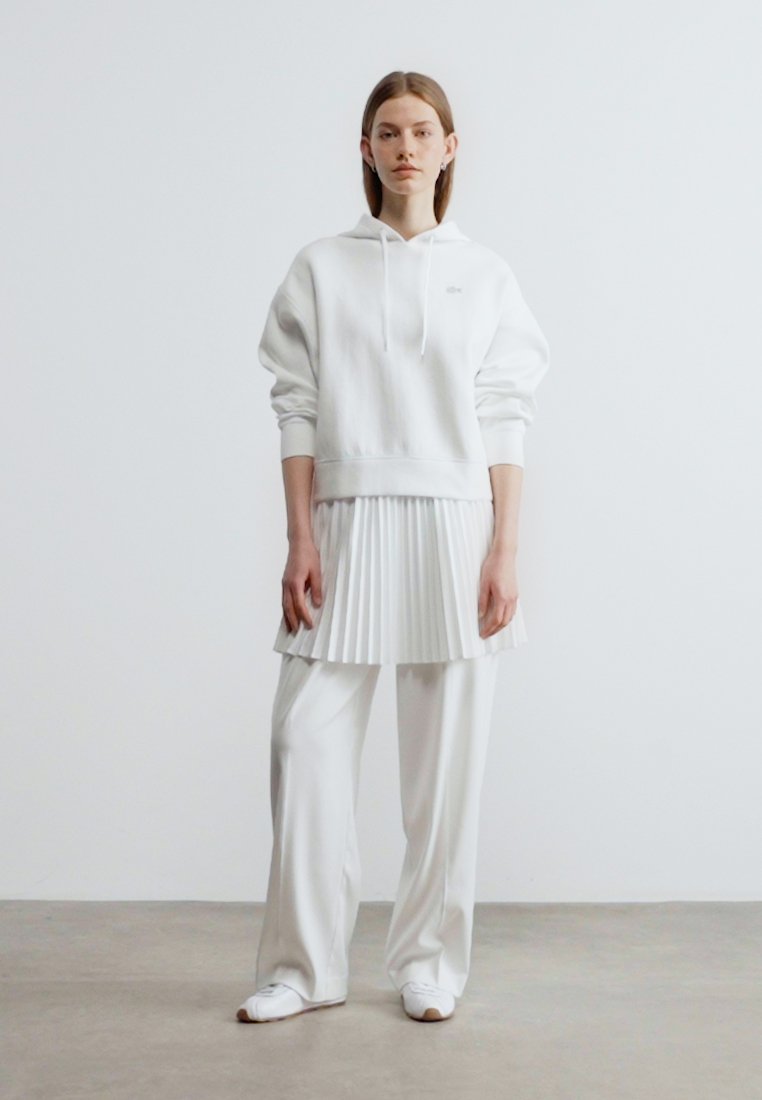 Femme debout portant un sweat à capuche blanc, une jupe plissée blanche sur un pantalon large blanc, et des baskets blanches, sur un fond uni.