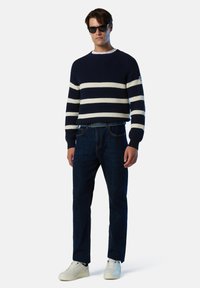 Jersey de punto a rayas en azul marino y blanco, jeans azul oscuro y zapatillas blancas. El modelo lleva gafas de sol, con tejido texturizado y detalles acanalados.