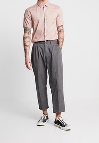 Chemise à manches courtes, rose clair, à boutons, avec un motif à pois ; pantalon gris, ample ; baskets noires avec accents blancs.