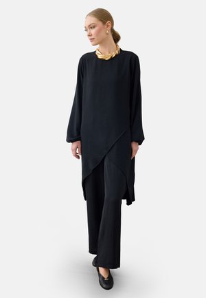 Frau trägt eine schwarze Tunika mit langen Ärmeln und asymmetrischem Saum, schwarze weit geschnittene Hosen, schwarze Schuhe und goldenen Schmuck, steht vor einem einfarbigen Hintergrund.