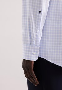 Camicia a quadri azzurro chiaro con una texture liscia, caratterizzata da un fiore ricamato in nero sulla manica e bottoni bianchi sul polsino.