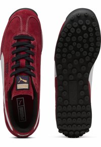 Sneakers roșii din suede cu șireturi negre, detalii albe pe lateral și o talpă din cauciuc texturat negru cu modele circulare. Logo-ul Puma pe limbă.