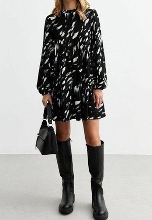 Kobieta ubrana w czarno-białą mini sukienkę z długim rękawem w wzory, czarne kozaki za kolano, trzymająca czarną torebkę.