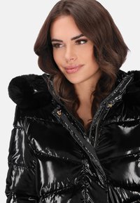 Fekete pufi dzseki fényes kivitelben, szőrmével bélelt kapucnival, cipzáras záródással és arany színű díszítéssel. Texturált felület steppelt mintával.