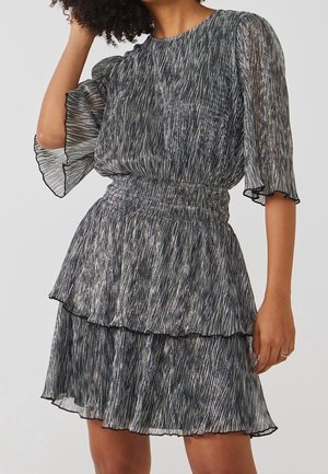 Frau trägt ein plissiertes, metallisch silber-schwarz gemustertes Kleid mit gepufften Ärmeln und tailliertem Schnitt.