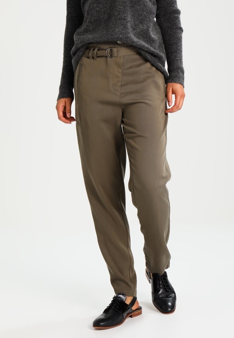 Pantalons vert olive clair avec une coupe décontractée, passants de ceinture et une texture lisse. Associés à des chaussures en cuir noires.
