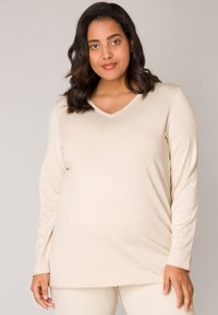 Beige Langarmshirt mit V-Ausschnitt, glatter Textur und lockerer Passform, das flache Nähte und einen leicht abgerundeten Saum aufweist.