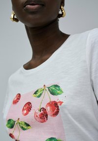 Camiseta blanca de manga corta con un gráfico de cereza rosa, hojas verdes y acentos rojos, que revela una tela de textura suave.
