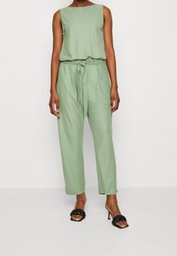 Ubit verde senza maniche con vita legata e pantaloni larghi; tessuto morbido e design minimal. Abbinato a sandali neri con tacco.