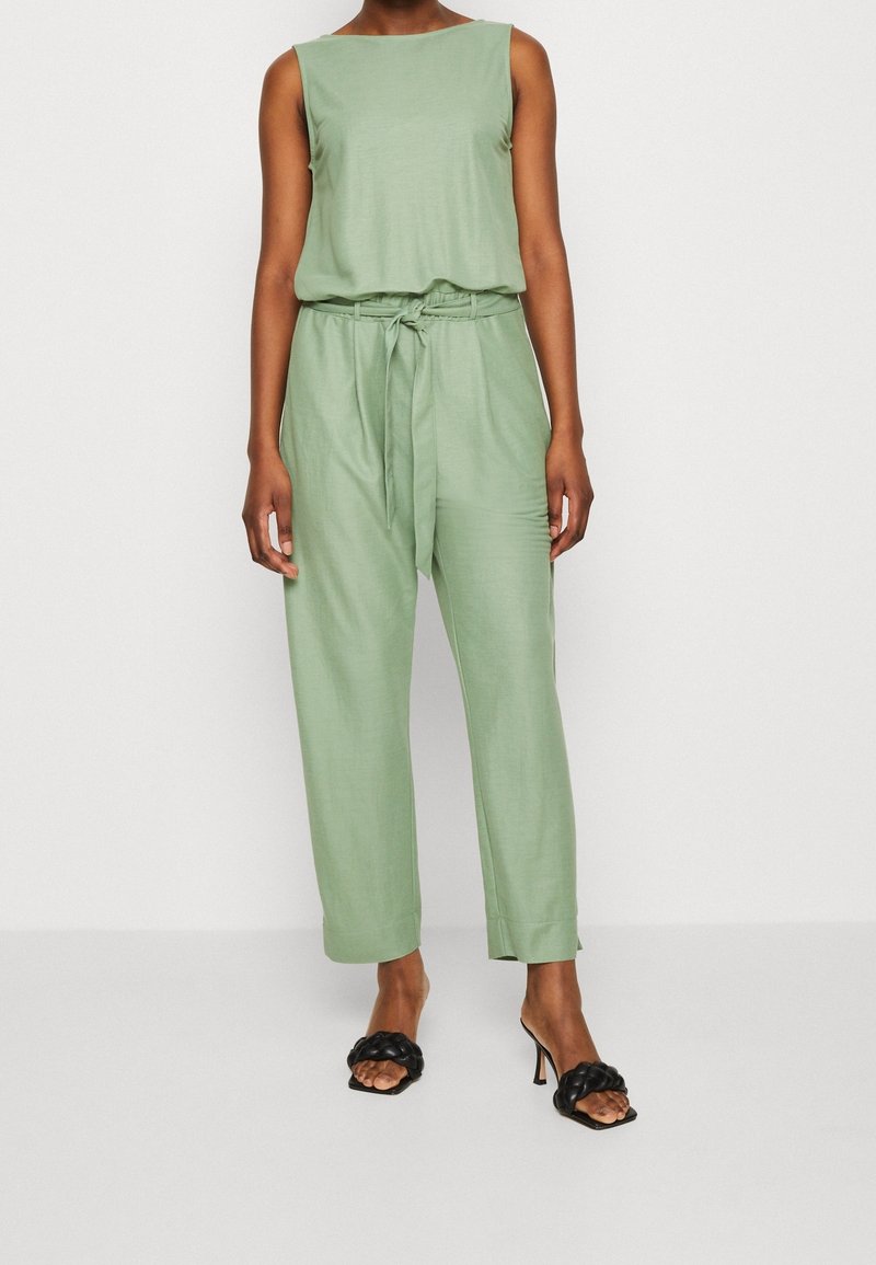 Ubit verde senza maniche con vita legata e pantaloni larghi; tessuto morbido e design minimal. Abbinato a sandali neri con tacco.