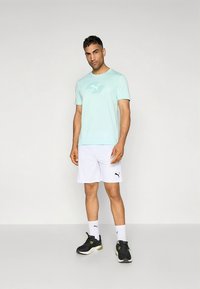 Lyseblå trenings-T-skjorte med grønn logo, kombinert med hvite shorts. Modellen har på seg svarte joggesko og hvite sokker, og står mot en nøytral bakgrunn.