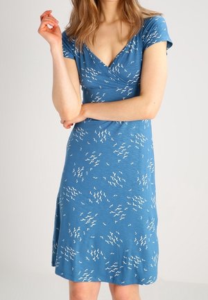 Kobieta ubrana w niebieską, krótkorękawową sukienkę kopertową z małymi białymi wzorami przypominającymi liście, stojąca z założonymi ramionami na tle jednolitym.