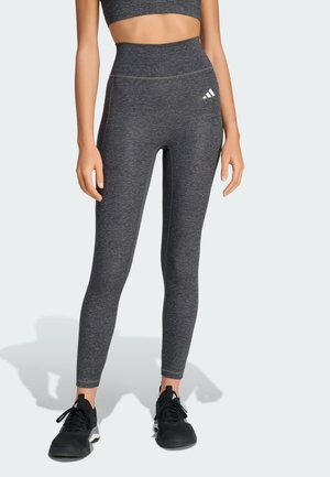 Žena nosící tmavě šedé legíny s vysokým pasem a odpovídající sportovní podprsenku s černými teniskami Adidas stojí na světlém pozadí.