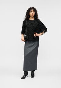 Haut noir à franges, jupe grise ajustée, bottines noires. Le mannequin tient un petit sac noir. Texture lisse et éléments de design en couches.