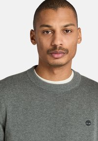 Grauer Pullover mit strukturiertem Strickmuster, Rundhalsausschnitt und kleinem schwarzen Logo auf der linken Brust. Darunter eine weiße Bluse.