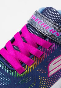 Navy színű hálós dizájnnal rendelkező Skechers cipők, élénk szivárványos kiegészítőkkel, rózsaszín fűzőkkel és fényvisszaverő elemekkel a pánton.
