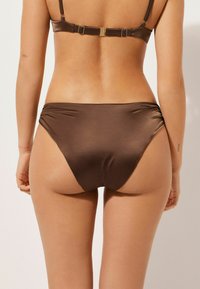 Bas de bikini marron avec une texture lisse et brillante. Présente des fronces sur les côtés pour un détail supplémentaire. Design simple avec une taille basse.