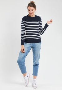 Maglione a righe blu navy e bianche con scollo a girocollo e dettaglio a bottoni sulla spalla sinistra, abbinato a jeans blu chiaro e sneakers bianche.