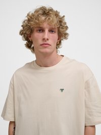 Jong persoon met krullend blond haar en gouden ringoorbellen, die een beige T-shirt draagt met een klein groen bijlogo op de borst.