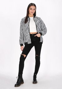 Černobílá bomber bunda se zebrovaným vzorem a ribbed manžetami, nošená přes bílý crop top, v kombinaci s černými roztrhanými skinny džíny a bojovými botami.