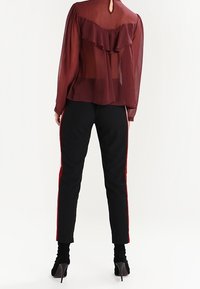 Blouse bordeaux transparente avec détail de volants au dos ; assortie à un pantalon noir ajusté avec des rayures rouges sur les côtés et des chaussures à talons hauts.