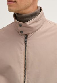 Lichtbeige jack met een hoge kraag, ritssluiting en knoopaccenten, gemaakt van een gladde, gestructureerde stof.