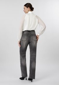 Γυναίκα με τα μαλλιά της σε κότσο, φοράει λευκό πουκάμισο με μακριά μανίκια, ξεθωριασμένο μαύρο τζιν με ψηλή μέση και μαύρα παπούτσια με τακούνι, στέκεται πλευρικά.