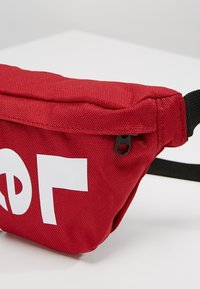 Sac banane en tissu rouge avec un curseur de fermeture éclair noir et un motif blanc partiel imprimé sur le devant, fixé à une sangle noire réglable.