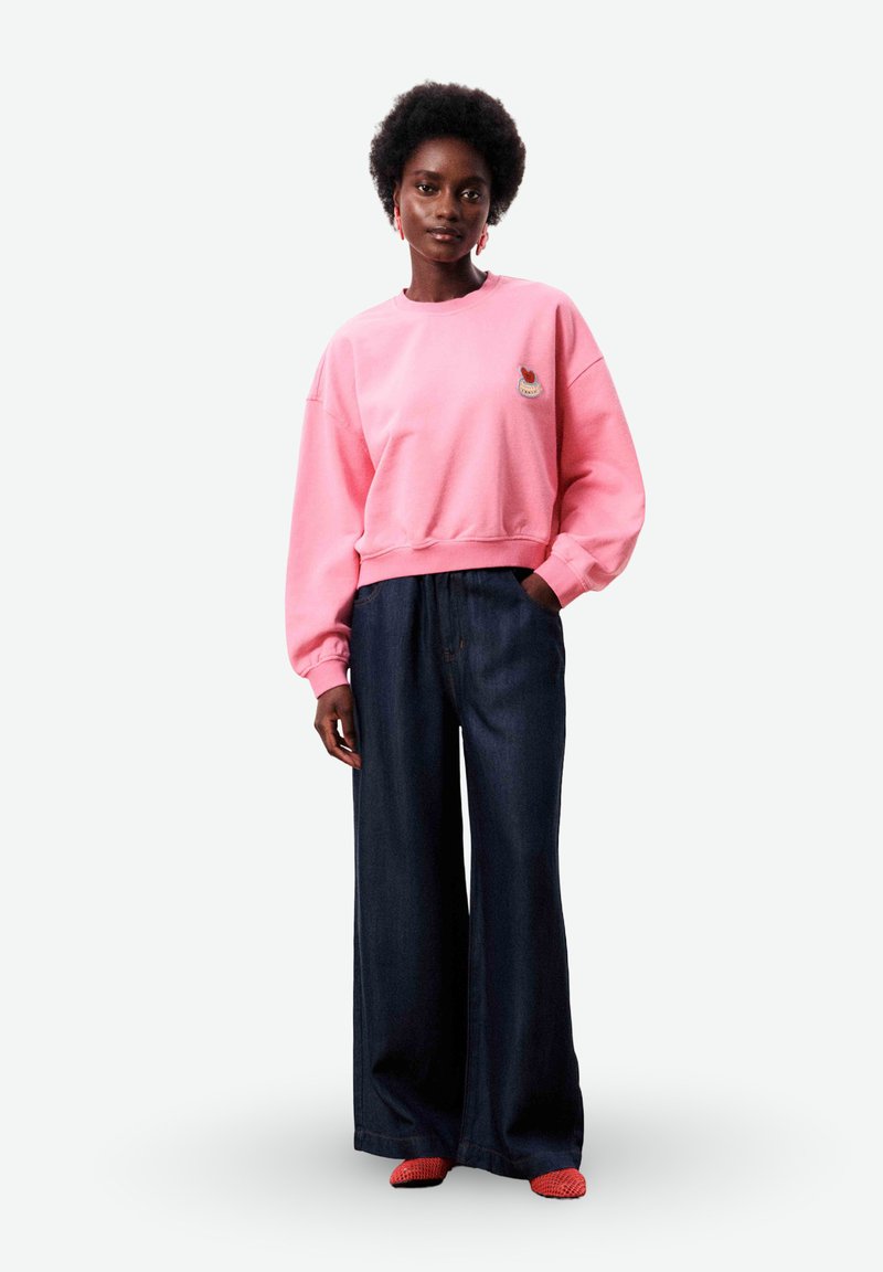 Woman with afro hairstyle wearing a pink sweatshirt with patch, wide dark blue pants, red earrings, and red shoes, standing with one hand in pocket.