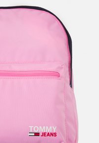 Zaino in tessuto rosa con una tasca frontale con zip, logo Tommy Jeans e un accento nero nella parte superiore. Superficie testurizzata con dettagli di cucitura.