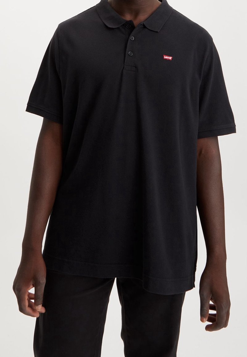 Personne portant un polo noir à manches courtes avec un petit logo rouge Levi's sur la poitrine gauche et un pantalon noir.