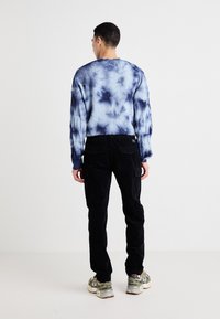 Pulover v temno modrem tie-dye vzorcu; dolgi rokavi, okroglo dekolte. Črne kordelyne hlače; sproščen fit s stranskimi cargo žepi. Bež in sivi superge.