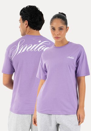 Mężczyzna i kobieta w lawendowych koszulkach z napisem „Smilodan”, stojący plecami do siebie na jednolitym białym tle.