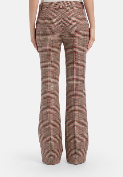 Person wearing high-waist plaid trousers in beige, red, and black, standing with hands relaxed at sides against a white background.