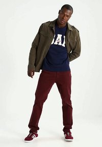 Mees, kes kannab oliivrohelist jopi, tumesinist GAP sviitrit, burgundi värvi pükse ja sellele sobivaid spordijalatooteid, seismas valge tausta ees.