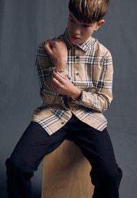 Beige kariertes Hemd zum Knöpfen mit blauen und schwarzen Akzenten, langen Ärmeln und einem lockeren Schnitt, kombiniert mit dunkler Jeans. Sitzt auf einem hölzernen Hocker.