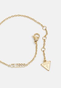 Zlatý náramok s logom "GUESS" a trojuholníkovým príveskom s vygravírovaným emblémom. Obsahuje sponu na homáre pre bezpečné uzatvorenie.