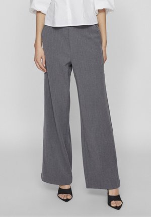 Vrouw die hoge taille, wijd uitlopende grijze broek draagt, gecombineerd met zwarte open-toe hakken en een witte blouse met pofmouwen.