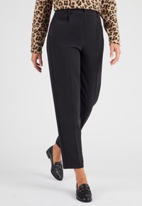 Zwarte op maat gemaakte broek met rechte pijpen, voorzien van voorplooien en een knoopsluiting. Draagt een gebreide trui met een patroon en zwarte instappers.