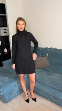Femme debout à côté d'un canapé en velours côtelé bleu, portant une robe noire en maille à col roulé et des talons noirs à bout pointu dans un salon.