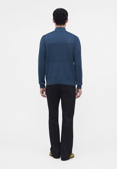 Homme debout, de dos, portant une chemise bleue à manches longues, un pantalon noir et des chaussures jaunes sur un fond blanc uni.