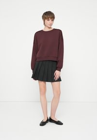 Bordový sveter s textúrovanými ramenami, skombinovaný s čiernou plisovanou sukňou. Modelka má na sebe čierne baleríny a stojí pred jednoduchým pozadím.