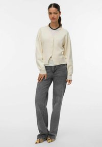Crème-kleurige gebreide cardigan met een V-hals, vier knopen en ingezakte schouders, gecombineerd met hoge grijze jeans en puntige schoenen met slangenprint.
