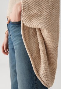 Gilet en tricot beige avec un motif texturé posé sur une poche de jeans en denim bleu, mettant en valeur un look décontracté et superposé.