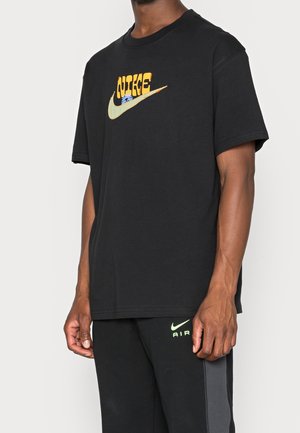 Mand iført sort Nike t-shirt med gult og beige logo og sorte Nike Air sweatpants med neongrønt logo.