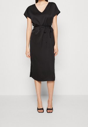 Kobieta ubrana w czarną satynową sukienkę midi z krótkimi rękawami, dekoltem w serek, paskiem przewiązanym w talii oraz czarne sandały na obcasie z odkrytymi palcami na białym tle.