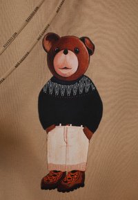 Gráfico de oso pardo en una camiseta color beige, vistiendo un suéter negro con un patrón geométrico blanco y pantalones color crema con detalles marrones.