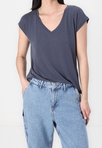 Žena v volném tmavě šedém tričku s výstřihem do V, zasunutém do světlých modrých džínů s pohodlným střihem, s rukama v kapsách.